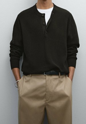 Homme portant un pull henley noir boutonné sur une chemise blanche, avec un pantalon plissé beige et une ceinture noire, les mains dans les poches devant un mur uni.