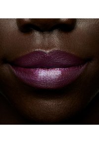 Gros plan de lèvres avec une couleur prune profonde, présentant une brillance et une texture lisse, sur fond de teintes de peau foncées. Un léger éclat est visible.