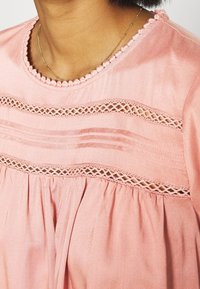 Blusa rosa con un motivo testurizzato, caratterizzata da inserti in pizzo decorativi e pieghe lungo l'orlo e il scollo. Tessuto morbido con una vestibilità rilassata.