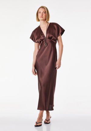 Kvinde iført en brun satin midi-kjole med korte ærmer, dyb V-hals, sort vedhængshalskæde og sorte sandaler med hæl på hvid baggrund.