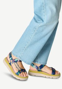 Sandali a fantasia floreale con un design di fasce multicolori, dotati di una suola beige imbottita e una base verde, indossati con jeans azzurri chiari.