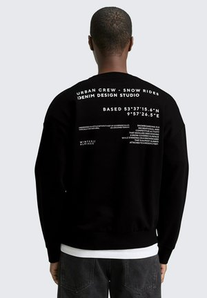 Persona vistiendo una sudadera negra con texto blanco en la espalda, que incluye "Urban Crew - Snow Rides Denim Design Studio" y coordenadas geográficas.