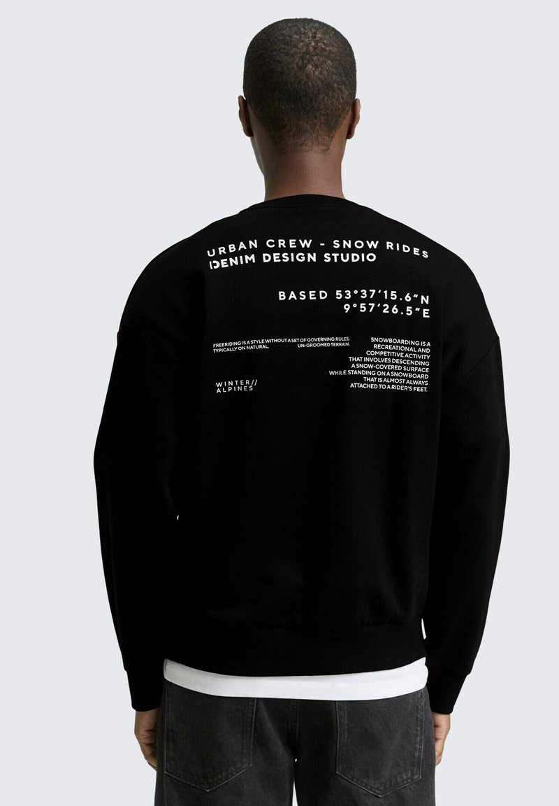 Personne portant un sweat-shirt noir avec du texte blanc au dos, incluant "Urban Crew - Snow Rides Denim Design Studio" et des coordonnées géographiques.
