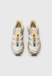 Αθλητικά παπούτσια σε ανοιχτό μπεζ χρώμα με καφέ λεπτομέρειες, υλικό από δίχτυ, αντανακλαστικές λεπτομέρειες και κίτρινη και μπλε ετικέτα στο πλάι.