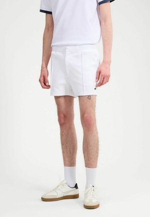 Mężczyzna w białych szortach, białych skarpetkach i białych tenisówkach z gumowymi podeszwami, stojący na tle jednolitego białego tła.