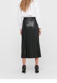 Jupe midi plissée noire en matière brillante, dotée d'une taille élastique et tombant sous les mollets. Portée avec des chaussures à talons hauts.