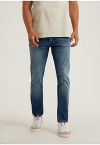 Modré džínové kalhoty s úzkým střihem, vybledlou texturou a klasickým pětipoklopovým designem. Kombinovány s bílými teniskami a světlou košilí.