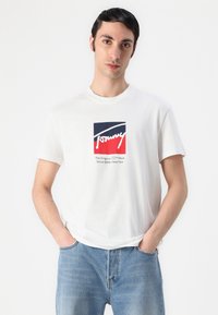Mladý muž s tmavými vlasmi, oblečený v bielej tričku Tommy Hilfiger a modrých džínsoch, stojaci s rukami v vreckách na jednofarebnom pozadí.