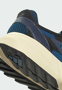 Sneaker met een blauwe mesh bovenkant, donkere grijze suède accenten, een witte tussenzool en een getextureerde zwarte rubberen buitenzool met drie grip patronen.