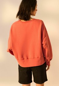 Koralowa, oversize bluza z opuszczonymi ramionami i prążkowanym dołem, w zestawieniu z czarnymi szortami dżinsowymi. Miękka faktura, minimalne detale.