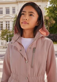Chaqueta con capucha de color rosa claro, con cintura ajustable mediante cordón, bolsillo con cremallera en el pecho, botones a presión y tela suave. Camisa blanca visible debajo.