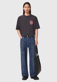 Czarny t-shirt z grafiką w okrągłym kształcie, w połączeniu z niebieskimi luźnymi dżinsami. Akcesoria to czarny pasek i teksturowana czarna torba na ramię.