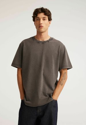Młody mężczyzna z kręconymi włosami i kolczykiem, ubrany w luźny szary t-shirt i ciemne spodnie, stojący z rękami w kieszeniach, pokazujący tatuaż na lewym przedramieniu.