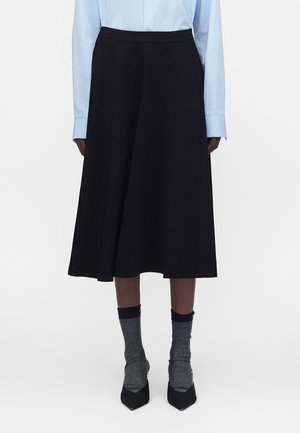 Personne portant une jupe noire mi-longue, une chemise bleu clair boutonnée, des chaussettes grises et des chaussures noires à bout pointu, debout devant un fond blanc.