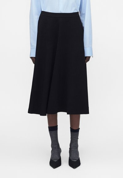Personne portant une jupe noire mi-longue, une chemise bleu clair boutonnée, des chaussettes grises et des chaussures noires à bout pointu, debout devant un fond blanc.