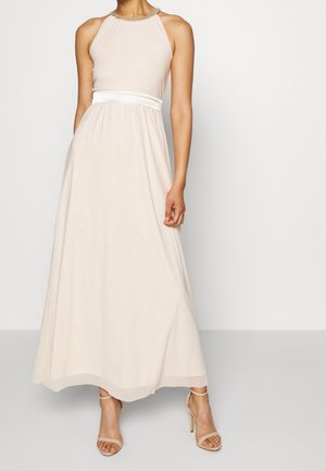 Kvinde iført en ærmeløs beige maxikjole med en hvid taljebånd og nøgen højhælede sandaler, stående mod en ensfarvet baggrund.