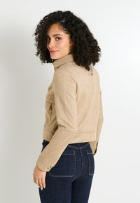 Veste en daim beige avec un col, une poche poitrine et des poignets boutonnés, design cropped, texture lisse, portée avec un jeans en denim foncé.