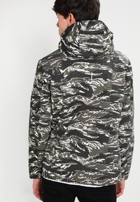 Grønn og hvit camouflert jakke med hette, laget av teksturert stoff og med sidelommer, sett fra baksiden.