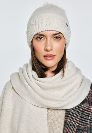 Jasnobeżowa dzianinowa czapka z warkoczowym wzorem i pomponem. Miękki, dopasowany beżowy szalik luźno owinięty wokół szyi. Struktura wydaje się przytulna.