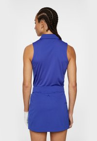 Blå ärmlös golftröja med krage i texturerad tyg, kombinerad med en matchande blå kjol, som har en minimal varumärkeslogotyp på baksidan.