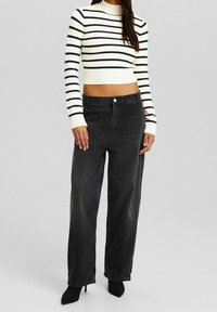 Svarta jeans med vid benkjol och hög midja, kombinerade med en krämfärgad och svart randig ribbad kortärmad tröja. Outfitten har figurnära ärmar och en hög krage.