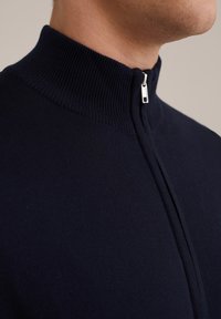Marinblå zip-tröja med hög ribbad krage och en mjuk yta, med en silverdragkedja och en figurnära design.