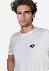 Weißes Baumwoll-T-Shirt mit einem kleinen, gestickten Sonnendesign in Dunkelblau und Gelb auf der linken Brustseite. Klassischer Rundhalsausschnitt.