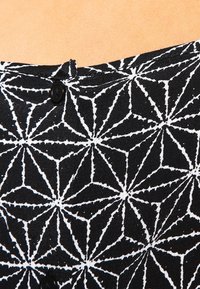 Svart tyg med ett geometriskt vitt mönster av korsande linjer. Två svarta knappar syns nära toppen.