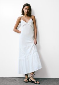 Robe blanche avec bord en dentelle et bretelles spaghetti, présentant un ourlet légèrement évasé et des coutures décoratives diagonales, accompagnée de sandales noires.