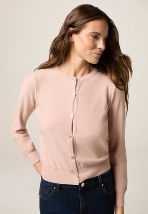 Femme aux longs cheveux bruns portant un cardigan à boutons rose clair avec des détails scintillants subtils et un jean bleu foncé, posant avec une main derrière le dos.