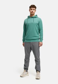Tyrkys hettegenser med teksturert stoff og snøring, kombinert med grå joggebukser. Modellen har på seg lyse sneakers og står på en ensfarget bakgrunn.