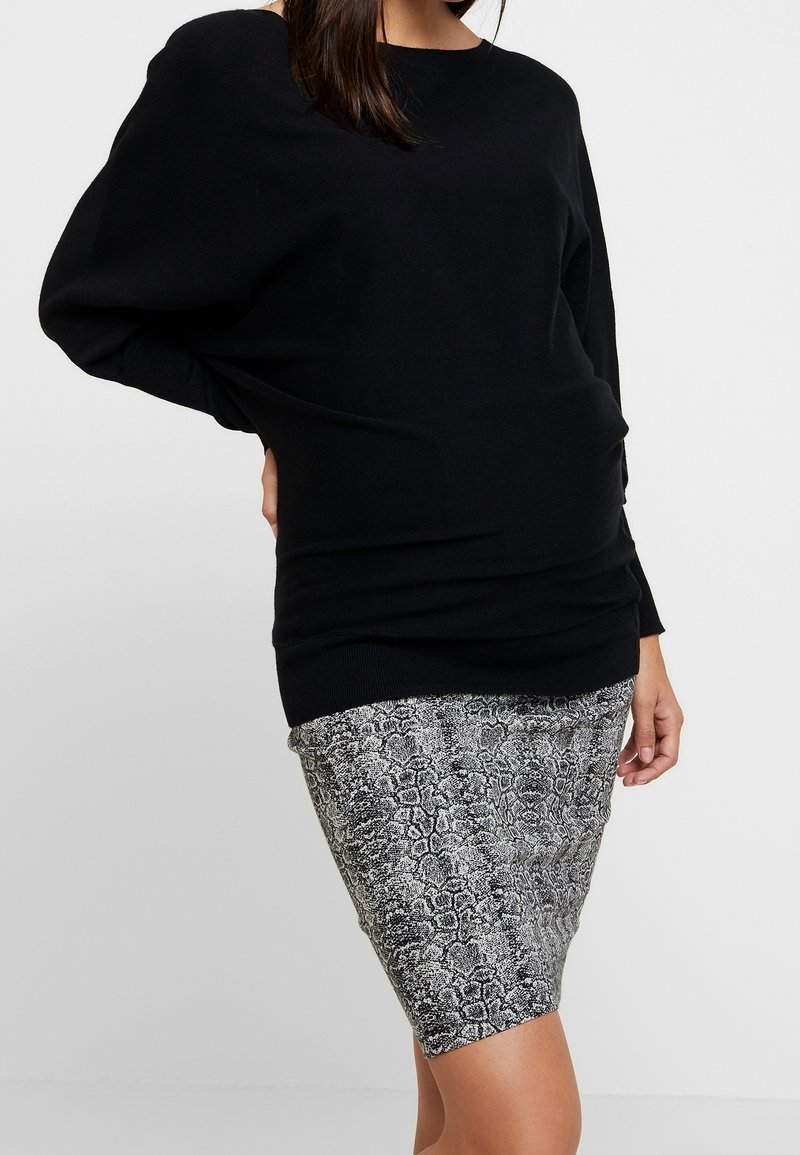 Femme portant un pull noir à manches longues et une jupe grise imprimé serpent jusqu'aux genoux, debout avec une main sur la hanche.