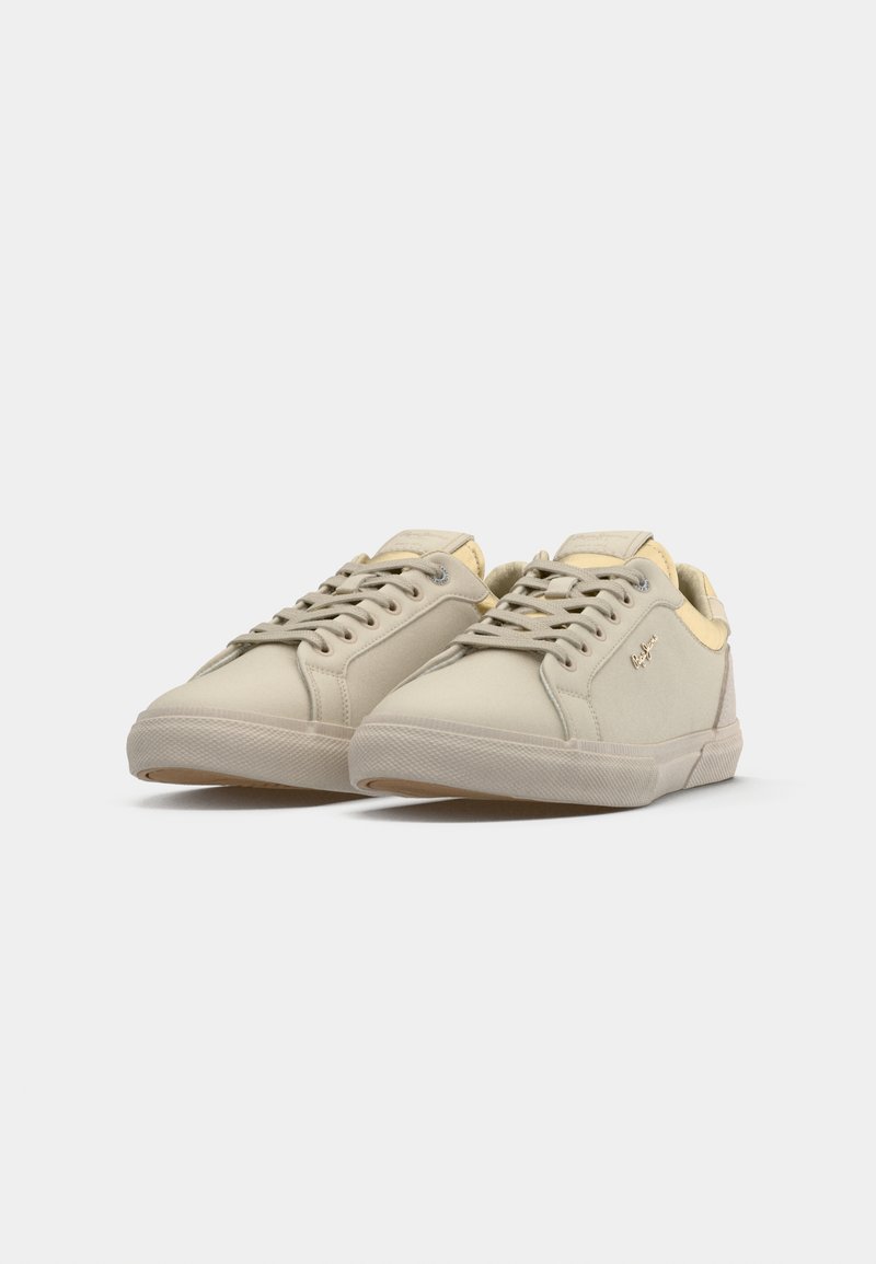 Zapatillas de lona beige con una textura suave, puntera redonda, cierre de cordones y sutiles acentos metálicos en los laterales. La suela de goma presenta un patrón de agarre.
