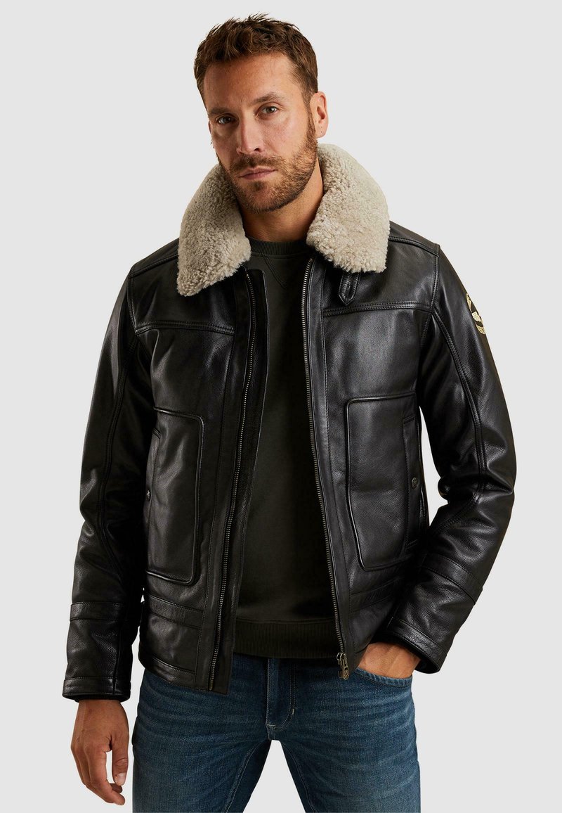 Črna usnjena jakna z bež ovčjim ovratnikom, z zadrgo in dvema sprednjima žepoma, oblečena čez zeleno majico s kapuco in modre kavbojke.