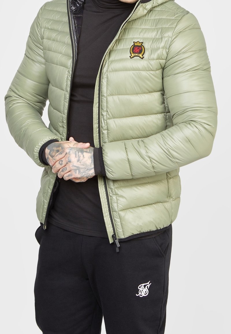 Man wearing light green quilted jacket with embroidered crest and black pants with white logo, hands clasped in front.