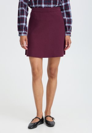 Donna con gonna a trapezio color bordeaux, camicia a maniche lunghe a quadri, anelli d'argento e scarpe Mary Jane nere, in piedi su sfondo bianco.