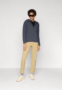 Mørkeblå zip-up jakke med hætte, teksturerede sidepaneler; parret med beige bukser og lyse sneakers. Model iført oversized sorte solbriller.