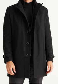 Cappotto nero realizzato in un tessuto liscio, caratterizzato da una chiusura con zip, dettagli con bottoni e un colletto alto. Maniche lunghe con polsini con bottoni.