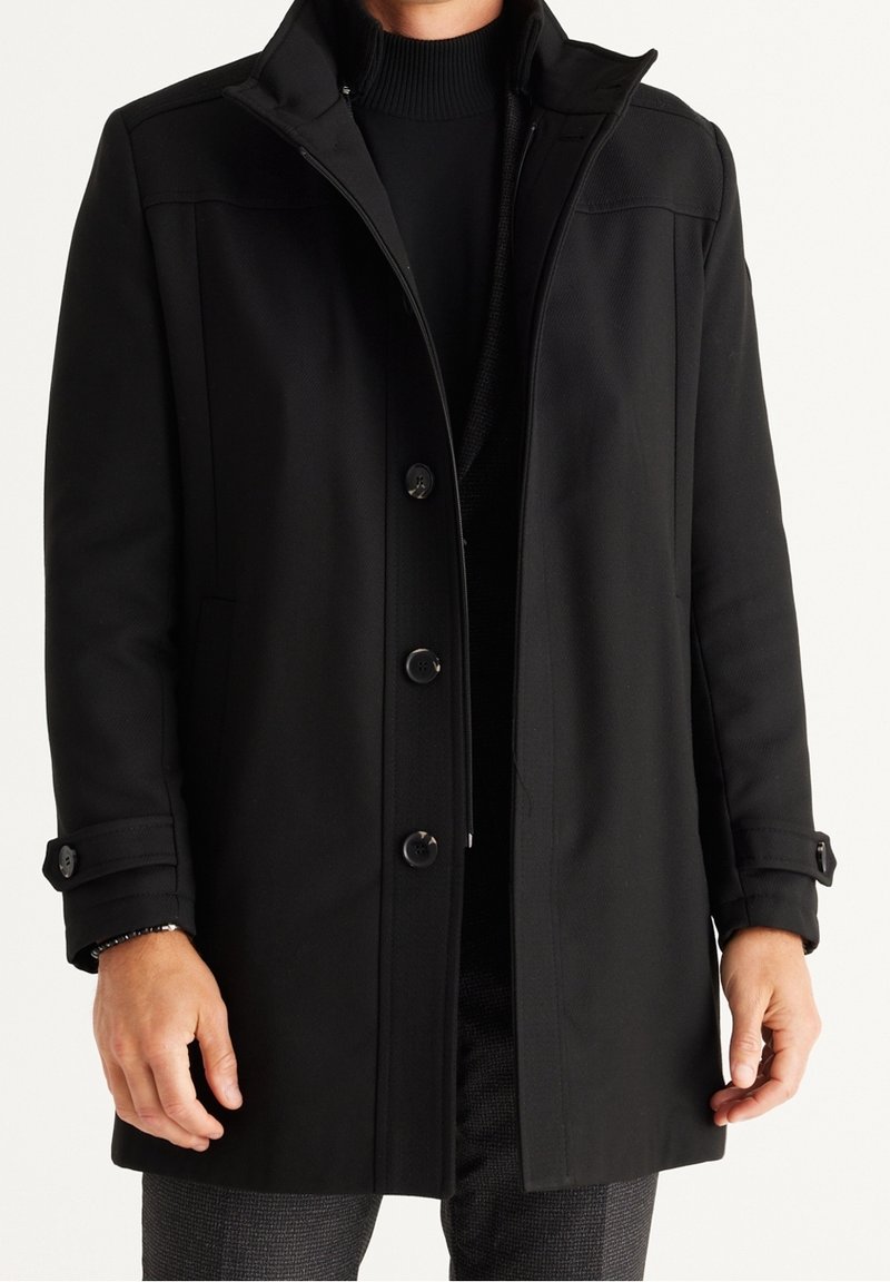 Cappotto nero realizzato in un tessuto liscio, caratterizzato da una chiusura con zip, dettagli con bottoni e un colletto alto. Maniche lunghe con polsini con bottoni.