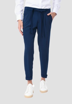 Persona che indossa pantaloni blu navy a gamba stretta con cintura annodata e risvolti arrotolati, abbinati a scarpe da ginnastica bianche e camicia bianca.