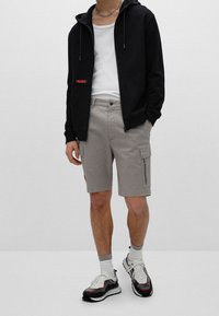 Hombre vestido con sudadera con cremallera negra, camiseta blanca, pantalones cortos de algodón grises, calcetines blancos y zapatillas negras y blancas, de pie contra un fondo liso.