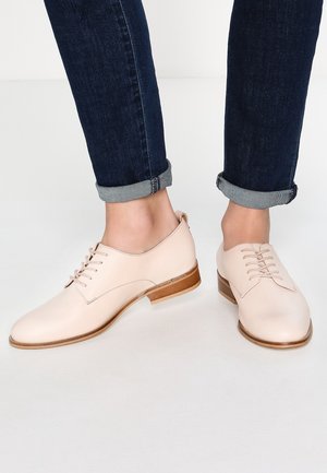 Jasnoróżowe skórzane oksfordy z okrągłym noskiem, sznurowanym zapięciem i drewnianym obcasem, noszone z podwiniętymi dżinsami.