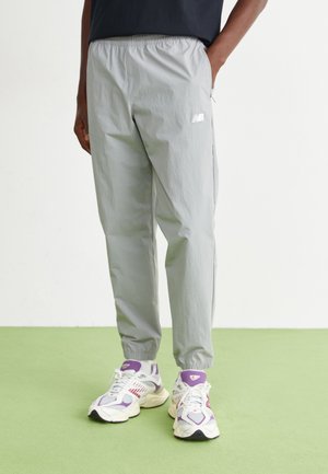 Unterkörper einer Person, die hellgraue New Balance Jogginghosen und weiß-violette New Balance Sneaker trägt und auf grünem Boden steht.
