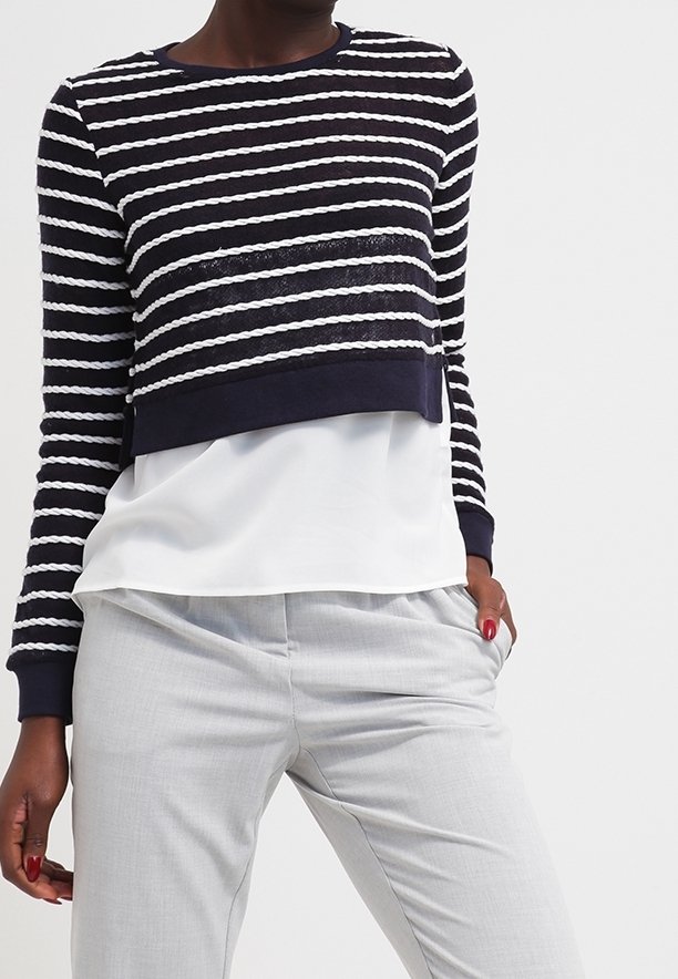 Personne portant un pull court à manches longues rayé noir et blanc par-dessus une chemise blanche ample et un pantalon gris clair, la main dans la poche.