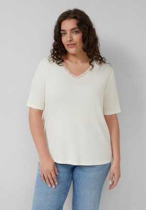 Kobieta z kręconymi brązowymi włosami, ubrana w biały, krótki rękaw z dekoltem w serek i koronkową lamówką oraz jasnoniebieskie dżinsy na szarym tle.