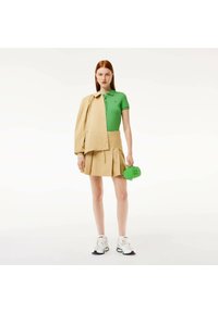 Groene polo, beige plooirok, beige jasje over de schouder, groene handtas en witte sneakers met zwarte accenten.