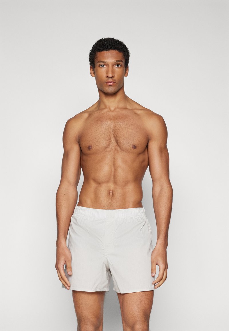 Jasnoszare szorty kąpielowe z elastycznym pasem, o prostym wzorze i długości do połowy uda, noszone przez męski model bez koszulki na neutralnym tle.