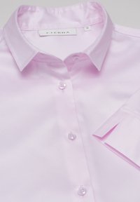 Camicia a maniche lunghe di colore rosa chiaro realizzata in tessuto morbido, con colletto a punta, sei bottoni e dettagli delle maniche piegate. Etichetta del marchio visibile.