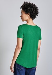 Zelené tričko s krátkým rukávem, mírně texturovaným povrchem, kulatým výstřihem a zakřiveným lemem, spojené s tmavě modrými džíny.