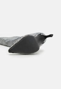 Zapato de tacón de aguja negro con parte superior texturizada adornada con tachuelas metálicas, punta afilada y suela de goma negra lisa.